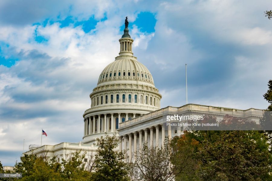 The US Capitol Building in Washington DC, at an angle, over some trees. The beautiful dome stretches up into the blue sky with scattered clouds.