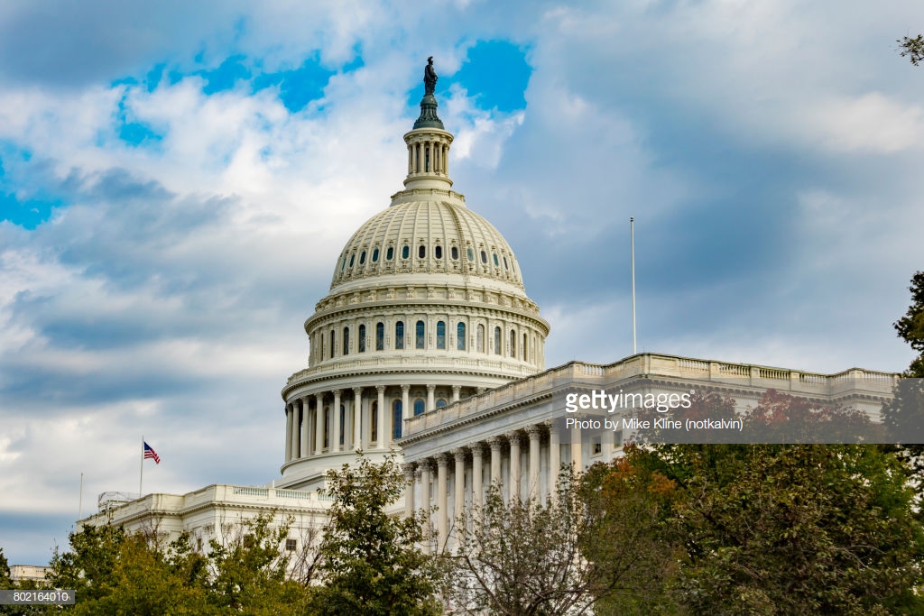 The US Capitol Building in Washington DC, at an angle, over some trees. The beautiful dome stretches up into the blue sky with scattered clouds.
